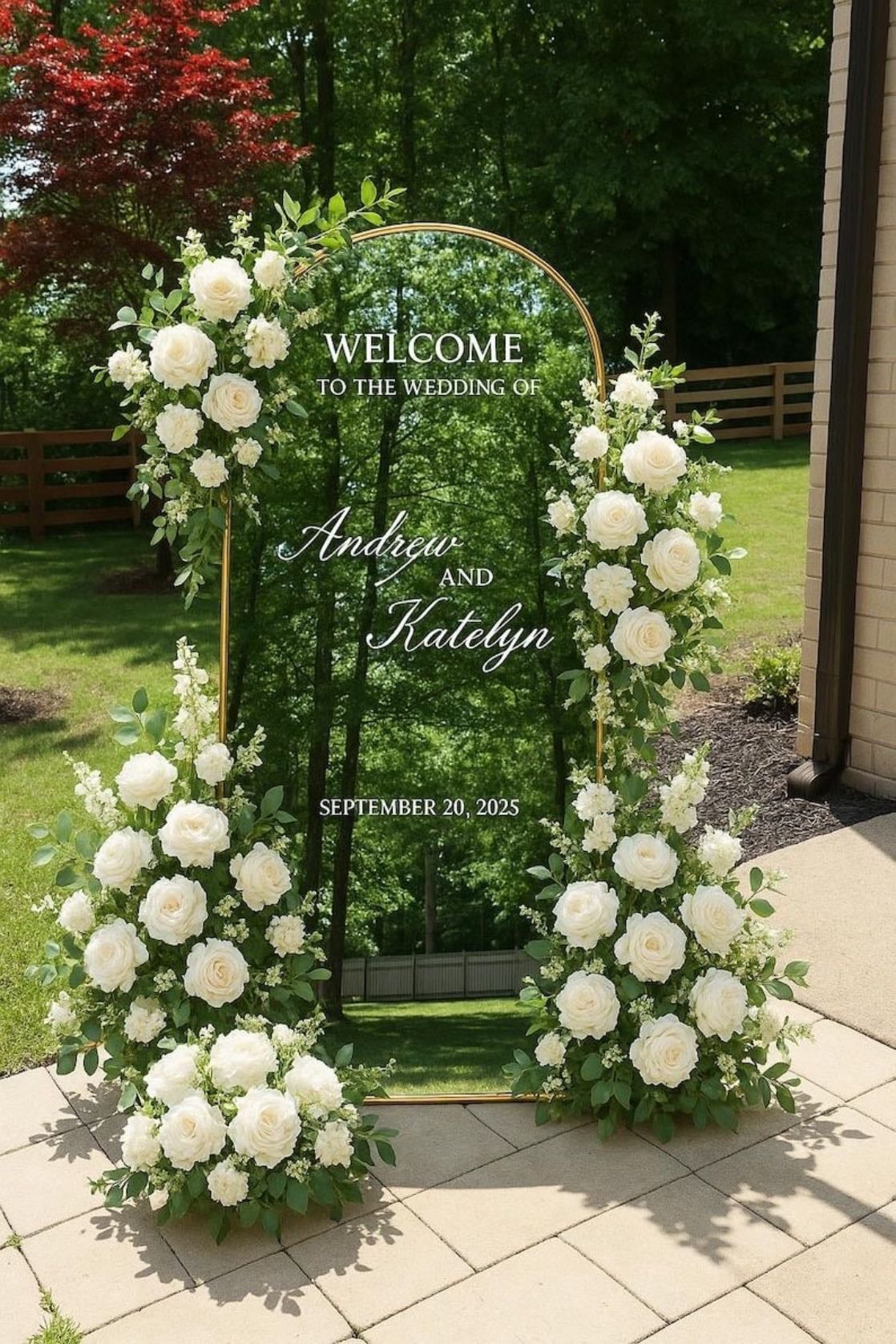 Espejo de bienvenida - Cartel de bienvenida elegante con base de espejo y estructura dorada, decorado con arreglos florales de rosas blancas y follaje verde. En el espejo, grabado el mensaje de bienvenida con los nombres de los novios y la fecha del evento, creando un detalle sofisticado y romántico para recibir a los invitados en la ceremonia.