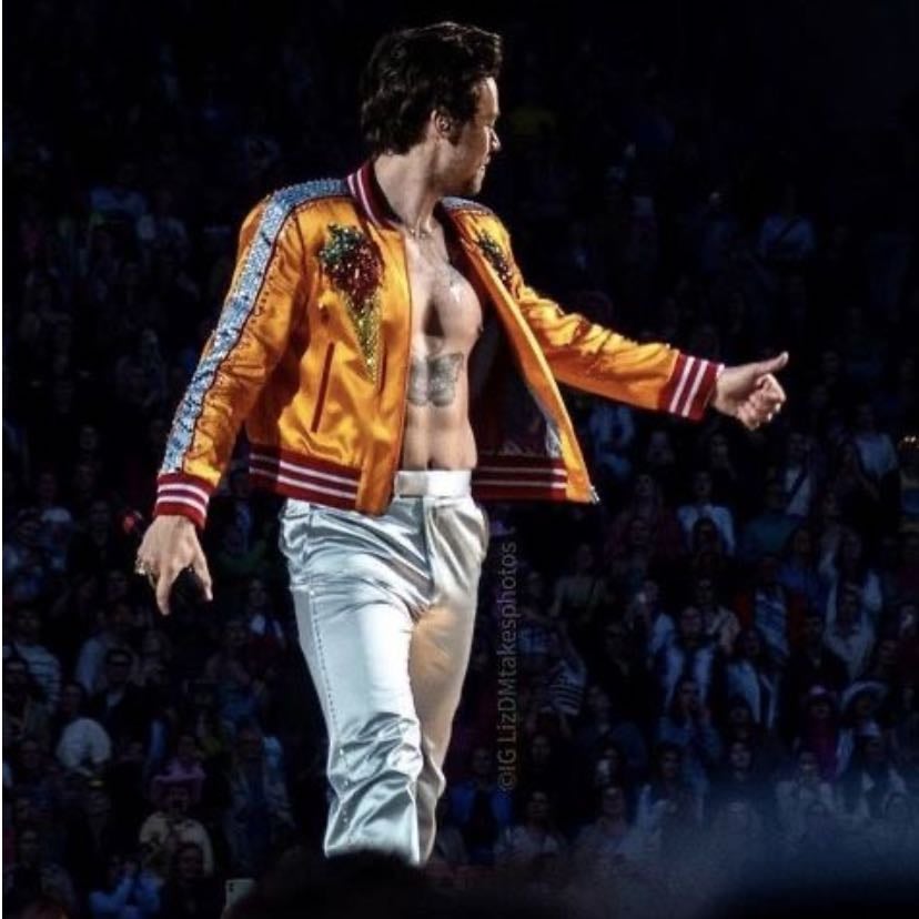 horsens 05/13/2023 - no shirt, orange and red letter jacket with rhinestones down the sides, a sparkly ice cream cone on the front, and a blue “Harry Styles” embroidered on the back, light blue satin pants, green satellite stompers